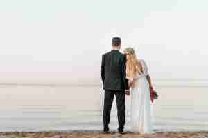 back view of wedding couple standing on beach with wedding bouquet and looking at sea